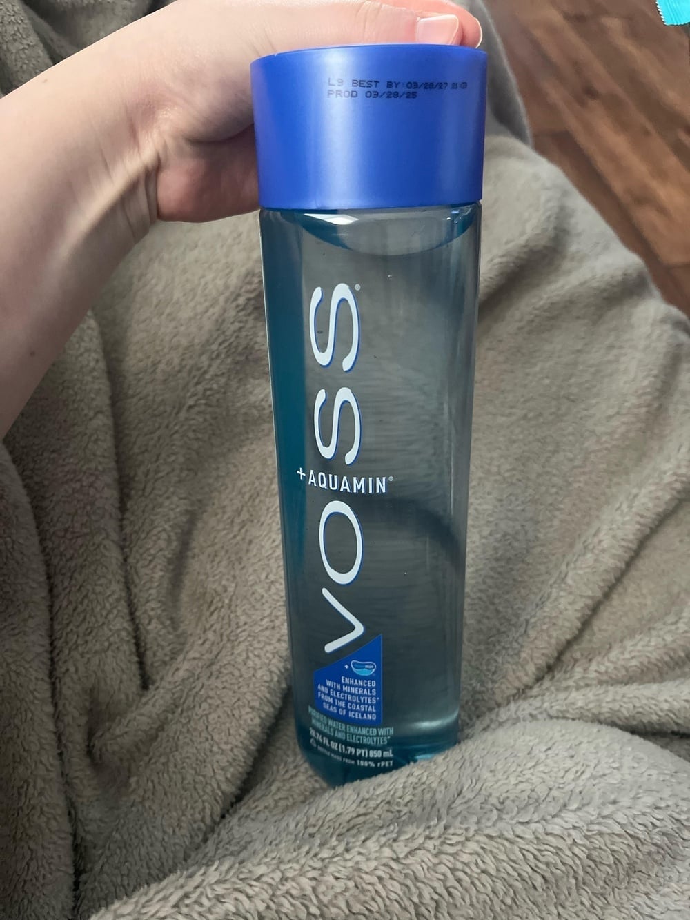 A hand holding a clear plastic bottle of Voss +Aquamin water with a blue cap.