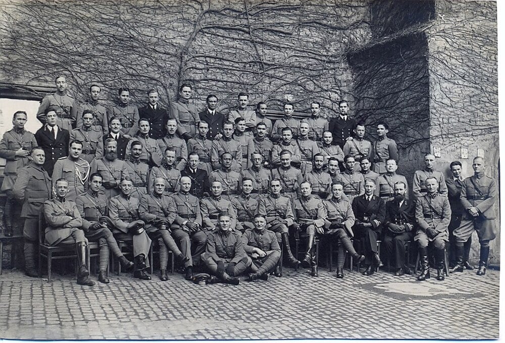 A large group of military officers in dress uniforms posed for a photograph in front of a stone wall covered in ivy.