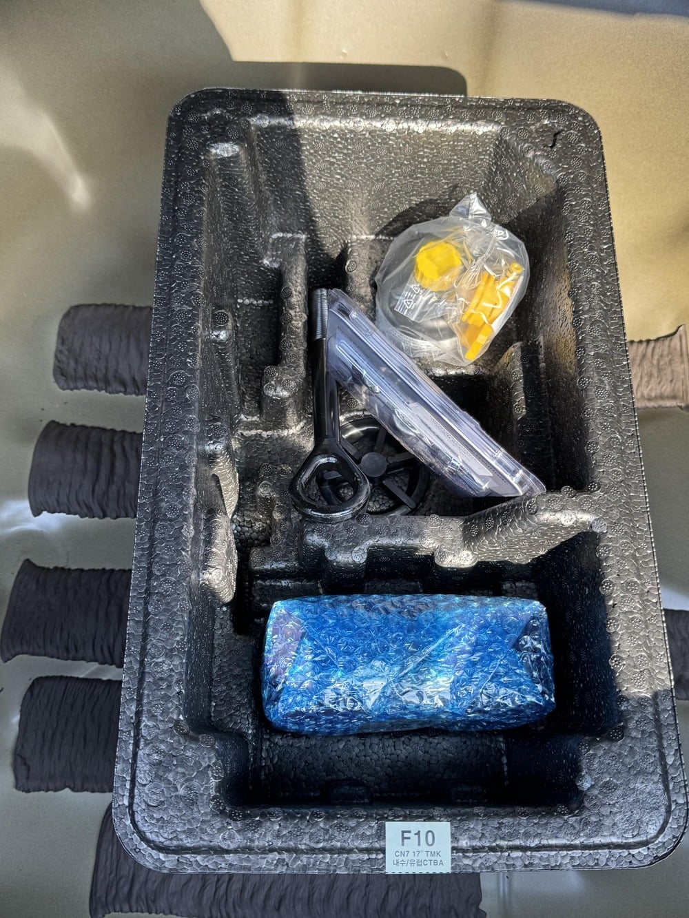 A black foam tray holds a plastic-wrapped blue item, a small clear container with yellow parts, and a black tool, all placed on a textured surface.