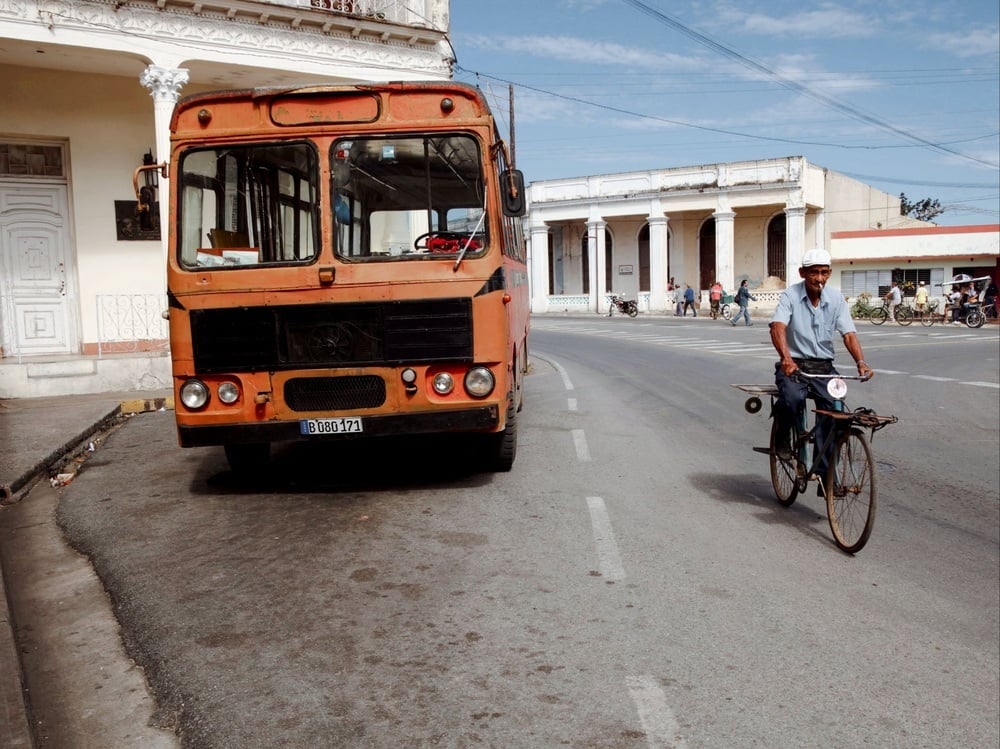 An older man rides a bicycle on a street next to a stationary orange bus.