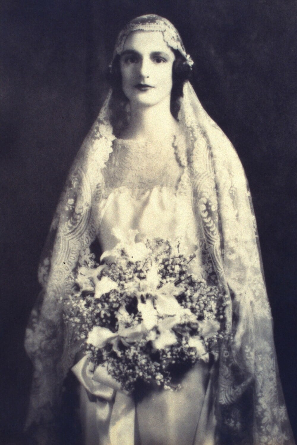 A vintage portrait of a bride wearing a lace veil over her head, holding a large floral bouquet.