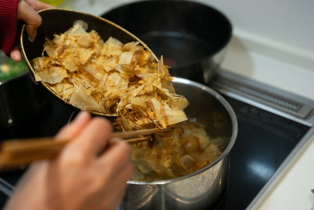 Hands use chopsticks to add dried bonito flakes from a bowl into a pot of simmering liquid on a stovetop.
