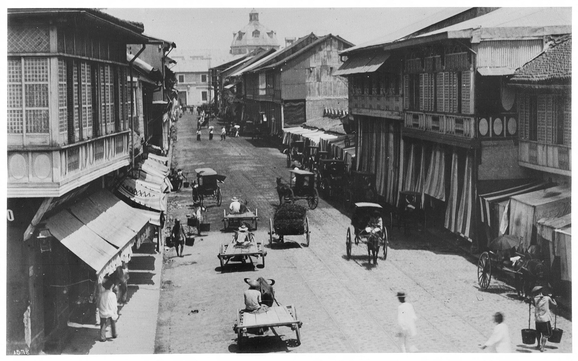 A black-and-white street scene in 19th-century Manila featuring wooden colonial houses with overhanging balconies, horse-drawn carriages, and vendors carrying goods on a busy thoroughfare.