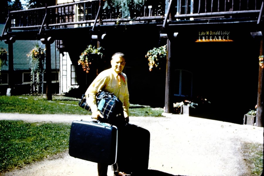 A man in a yellow shirt carries three pieces of luggage outside the Lake McDonald Lodge entrance.