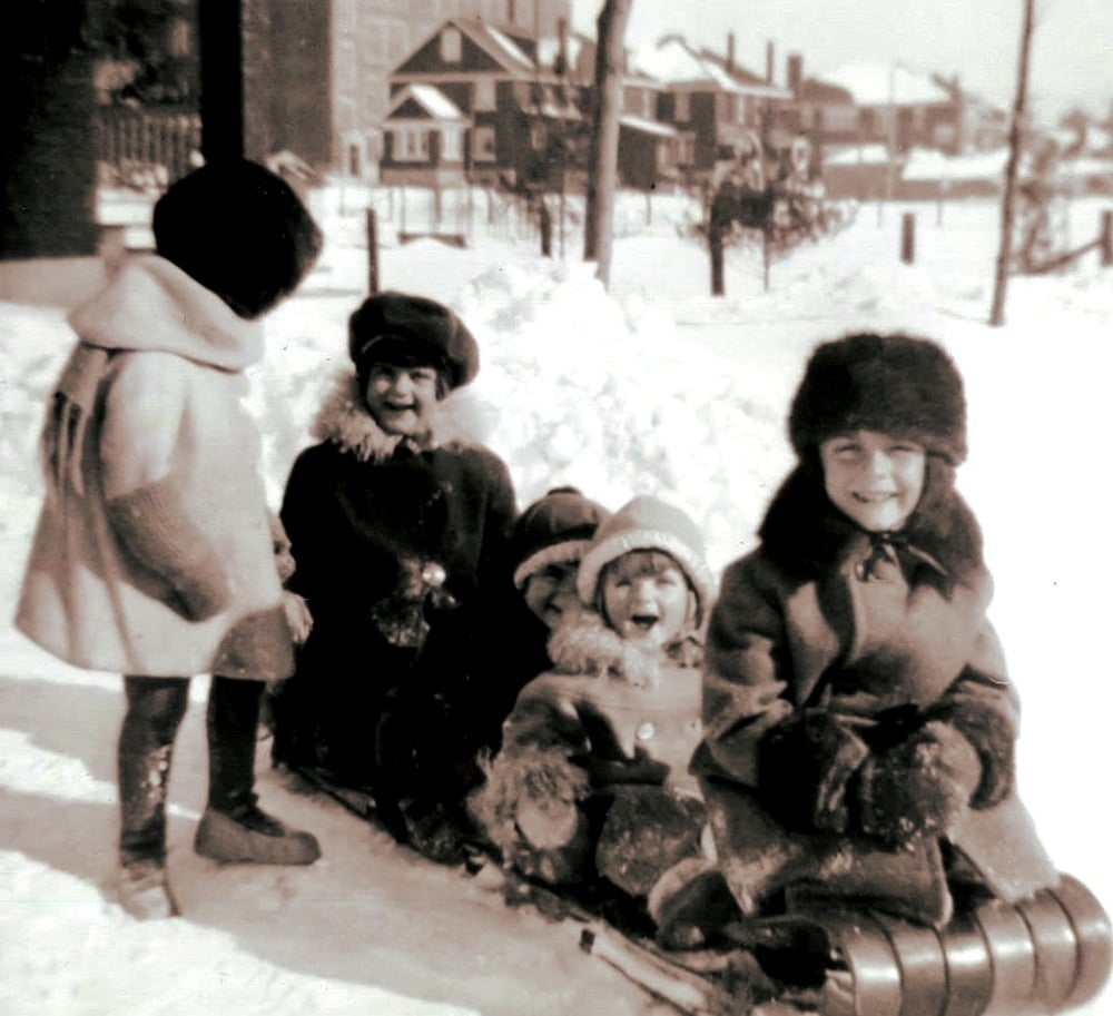 Four children in winter coats and hats sit on a sled in the snow, with another child standing beside them.