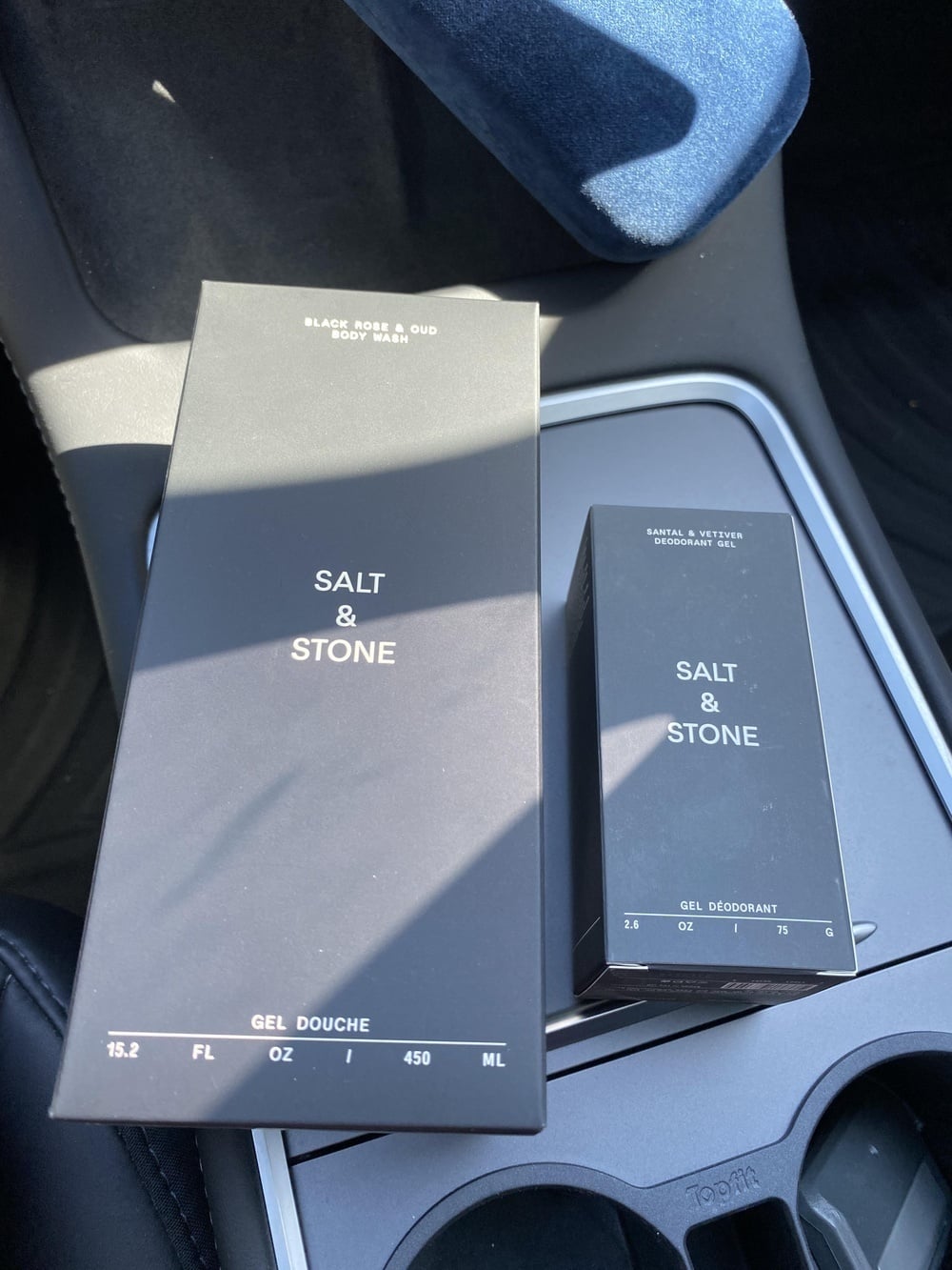Two Salt & Stone product boxes resting on a car console: a large Black Rose & Oud body wash and a smaller Santal & Vetiver deodorant gel.