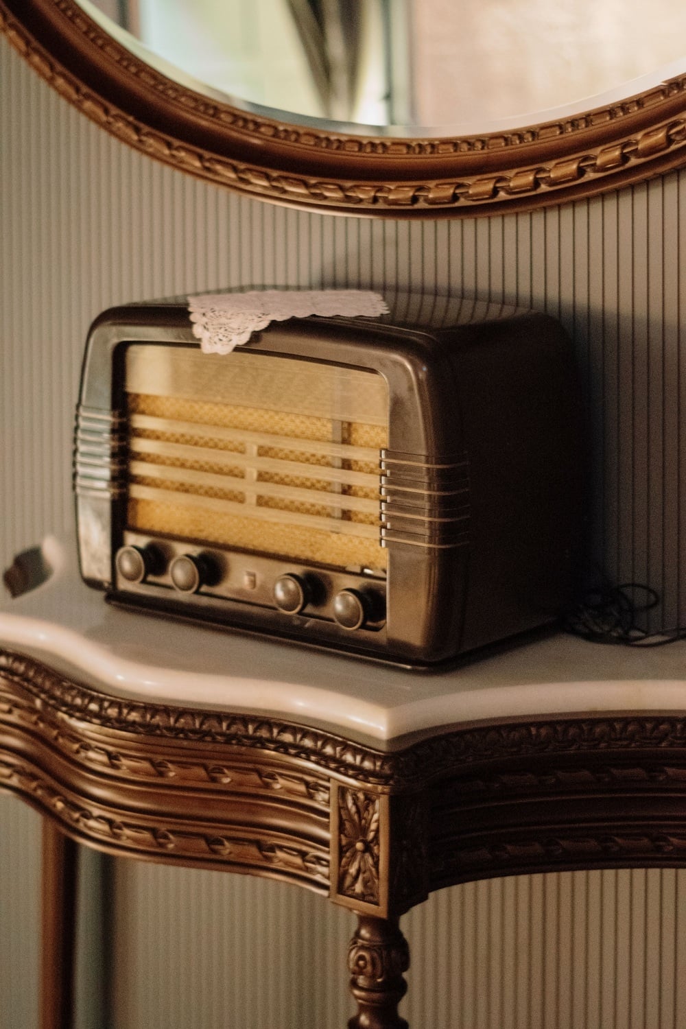 A vintage tabletop radio with a small lace doily on top, sitting on a wooden console table beneath an oval mirror.