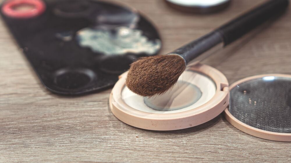 A makeup brush resting on an open compact of face powder.