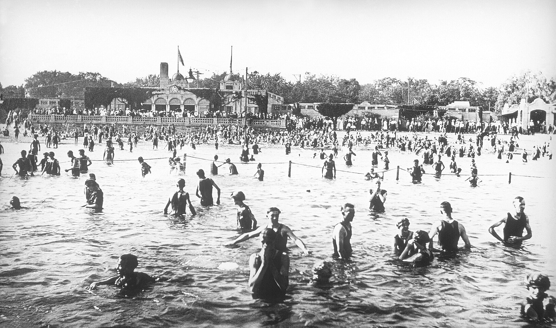 A black-and-white historical photograph of a crowded public beach with many people swimming in the water and a large pavilion visible in the background.
