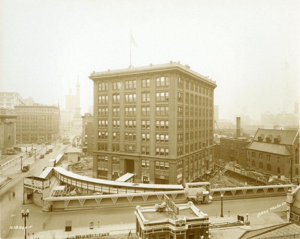 A high-angle, sepia-toned photograph of a large, multi-story office building surrounded by construction, with a pedestrian bridge crossing over a street in the foreground.