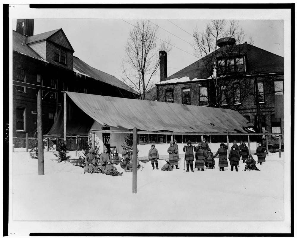 A group of people wearing heavy winter coats stand and sit in the snow in front of a long, low tent structure situated between two large brick buildings.
