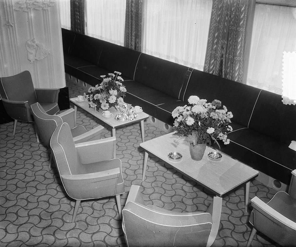 Interior of a passenger train car with upholstered armchairs, long bench seating, and two small tables topped with flower arrangements.