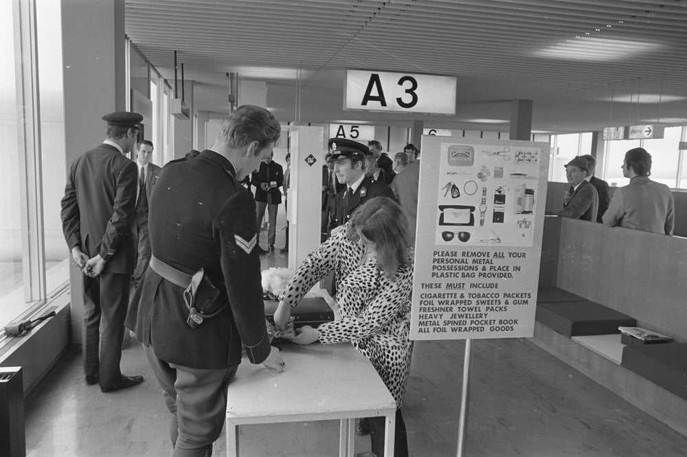 Black-and-white photo of airport security, 1960s. Officers check a woman’s bag at a table near a sign with inspection instructions. Other people wait in the background near a gate marked A3.