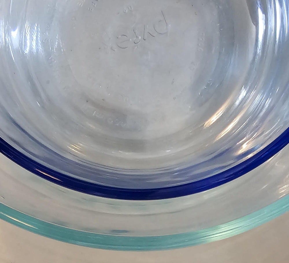 A close-up view of the bottom of a clear, round Pyrex glass bowl sitting on a light surface, with reflections and blue-green edges visible.