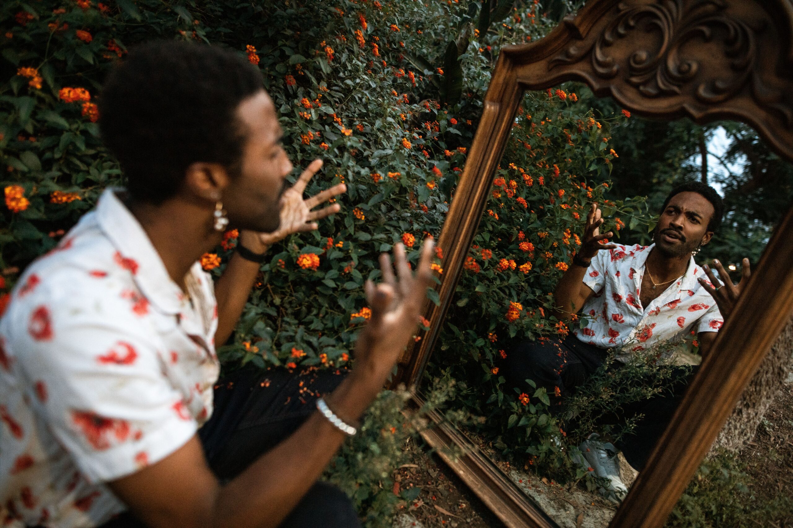 A person in a floral shirt gestures expressively while kneeling in front of a large ornate mirror outdoors, surrounded by orange flowers and greenery.