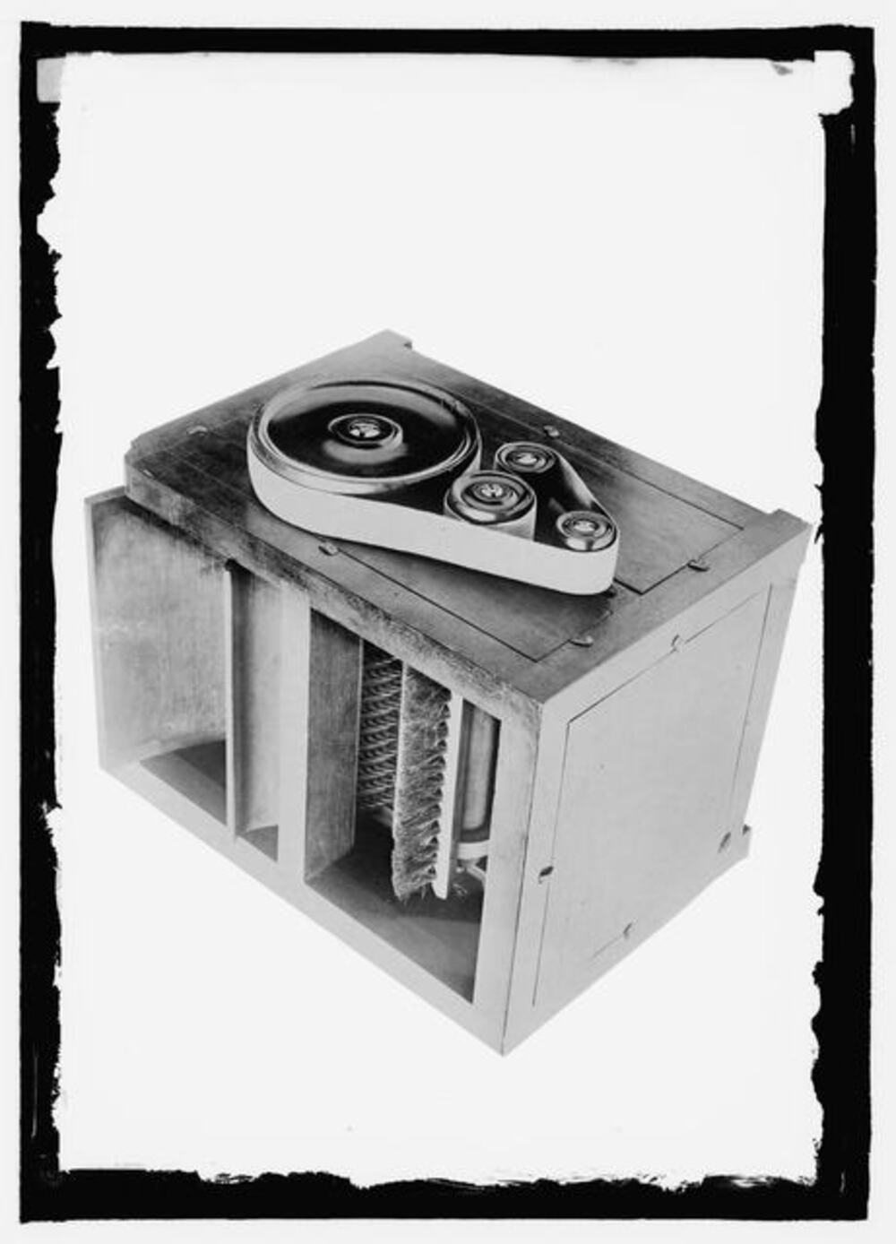 A wooden box-like machine featuring a belt-driven mechanism on top and a rotating brush cylinder exposed within the base.