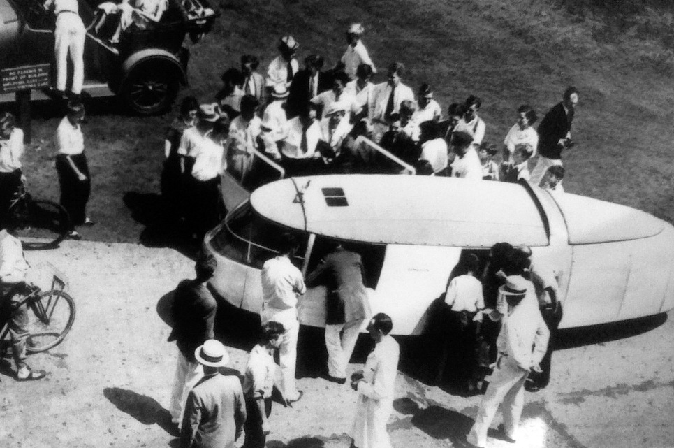 A large group of people gathers around a teardrop-shaped Dymaxion car in an outdoor lot.
