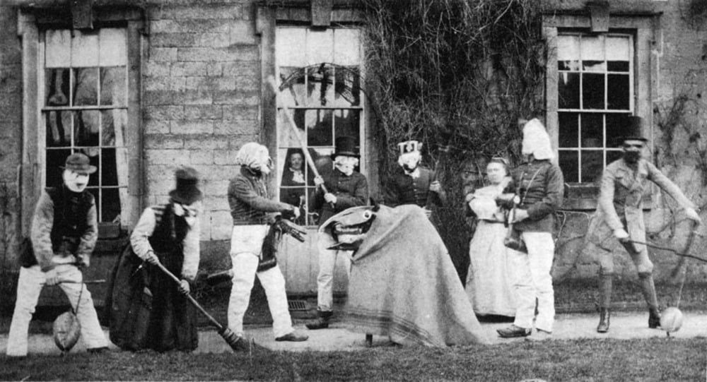 A group of people in costumes and masks standing in front of a stone building, with one person in the center wearing a pointed, draped head covering.