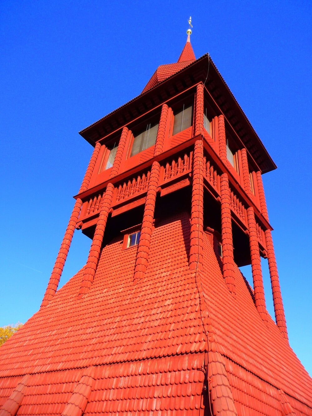 Tall, red wooden bell tower with shingled walls and an open gallery under a clear blue sky.