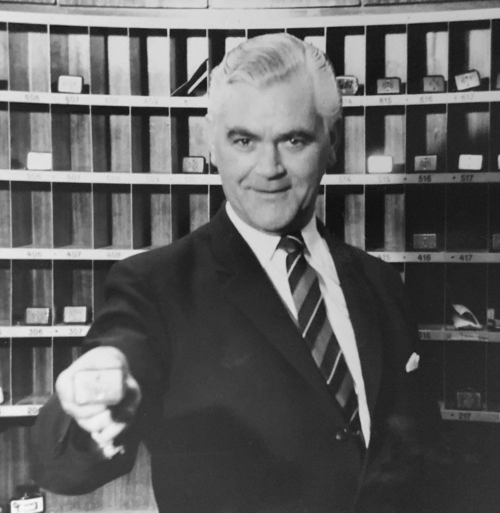 A man in a suit and striped tie standing in front of a hotel mail slot wall, holding out a room key.