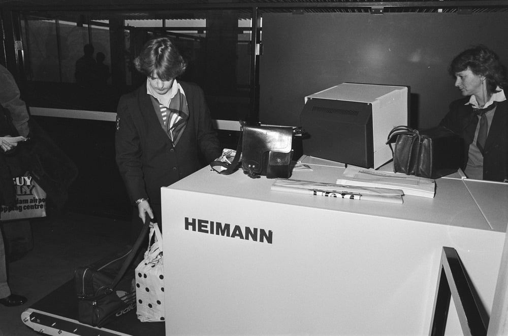 Two women at an airport security checkpoint place bags on a conveyor belt scanner labeled “HEIMANN.” One woman is looking down; the other holds a purse, preparing for screening.