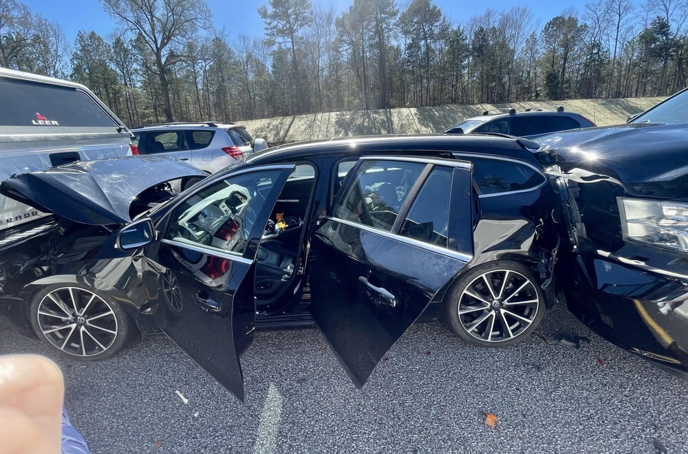 A black station wagon severely damaged in a multi-vehicle collision with its doors and hood open.