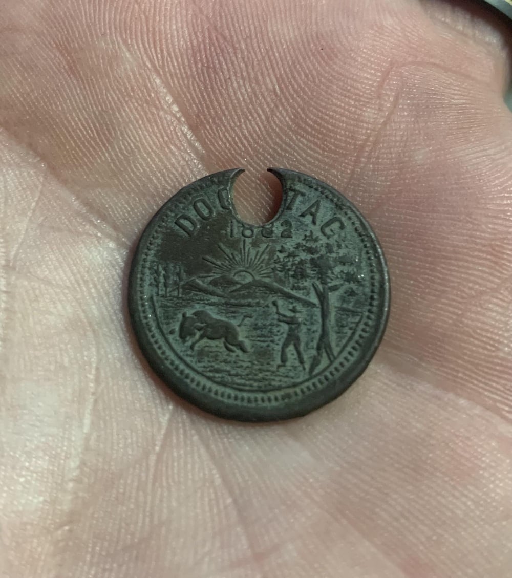 A metal dog tag dated 1882 held in a person’s palm, featuring an engraved scene of a person and an animal below a sunburst over mountains.