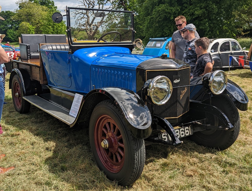 A blue and black vintage truck with wooden bed sides and red wheels parked on a grassy field.