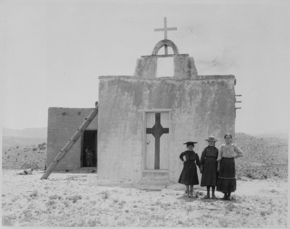 Three girls stand in front of a small adobe church with a cross on the door and a bell gable on the roof.