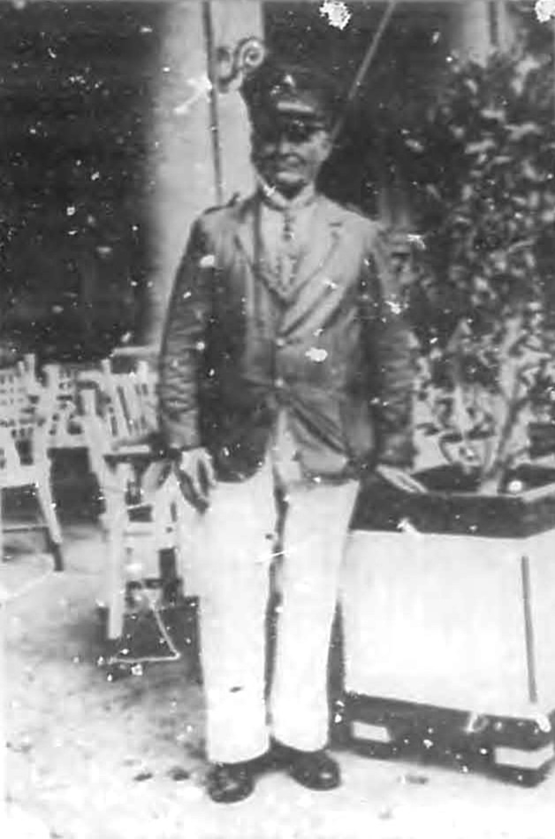 A man in a military-style uniform and cap standing outdoors near a potted plant and chairs.