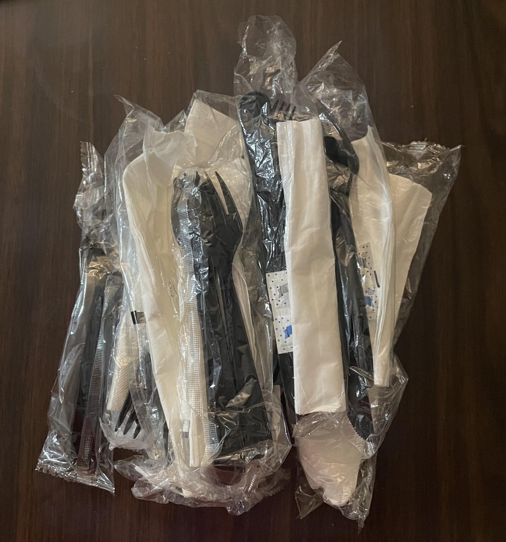 A pile of individually wrapped plastic cutlery sets, each containing a black fork, knife, and white napkin.
