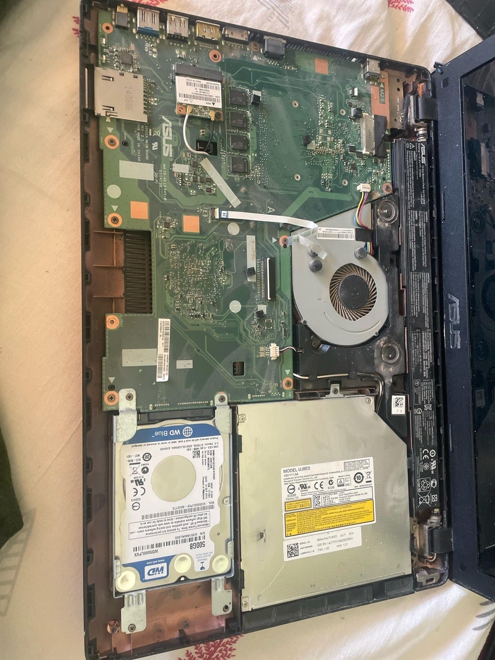 An open ASUS laptop showing internal components, including the motherboard, cooling fan, battery, hard drive, and optical drive, all placed on a white surface.