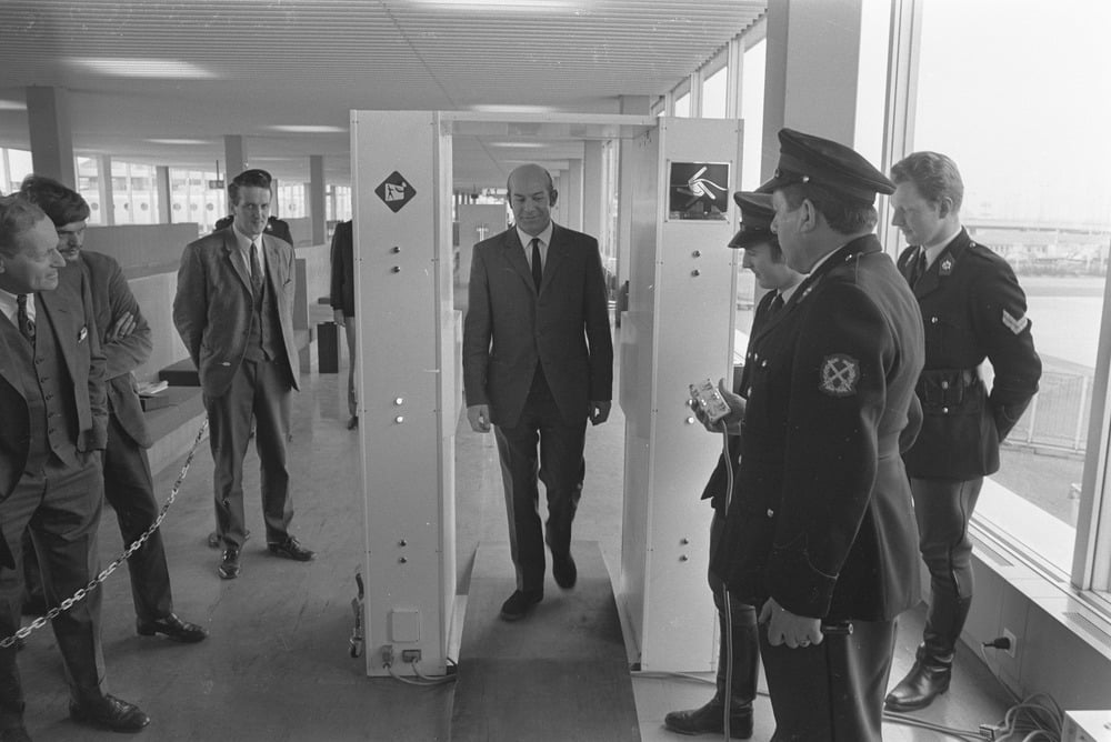 A man in a suit walks through a security metal detector, observed by several uniformed officers and other men in suits inside a bright building.