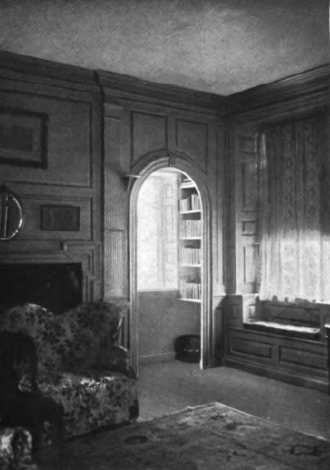 A paneled room with a patterned armchair, a window seat with curtains, and an arched doorway leading to a built-in bookshelf.