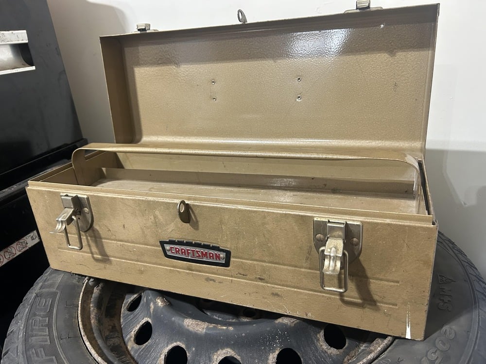 An open tan Craftsman metal toolbox with dual latches sits on a worn black tire. The box is empty and its lid is upright, showing the interior and the Craftsman logo on the front.