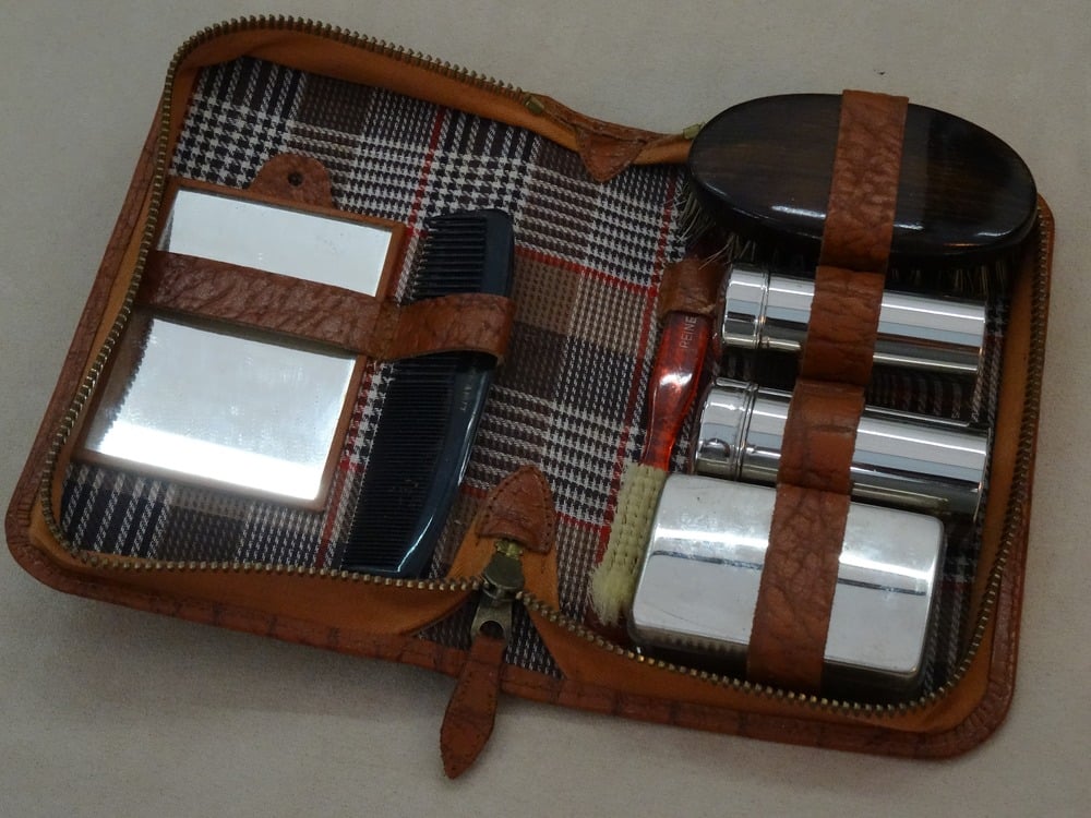 A vintage grooming kit with a brown leather case open to reveal a mirror, comb, brush, and metal containers, all neatly secured against a plaid fabric lining.