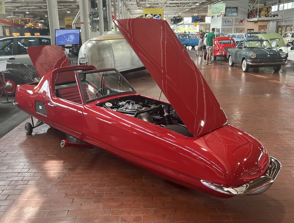 A red, teardrop-shaped prototype vehicle with its front hood open, displayed on a brick floor in an automotive museum.