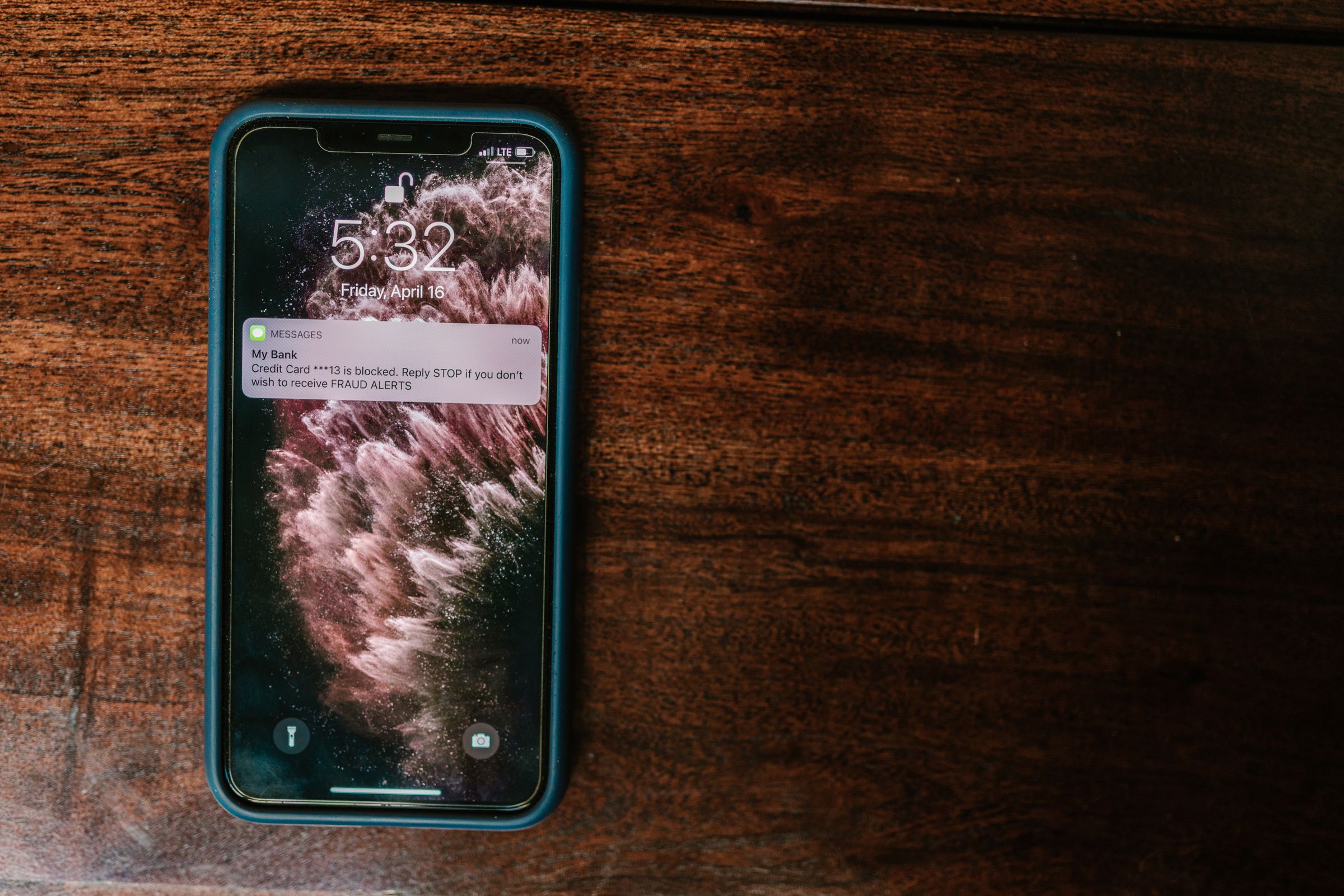 A smartphone on a wooden surface shows a notification about a blocked debit card due to fraud alerts. The time displayed is 5:32, and the background features a swirling, abstract pink and black pattern.