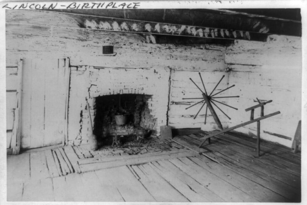 Black and white photo of the interior of a log cabin with a stone fireplace and a spinning wheel, labeled “Lincoln-Birthplace” at the top.