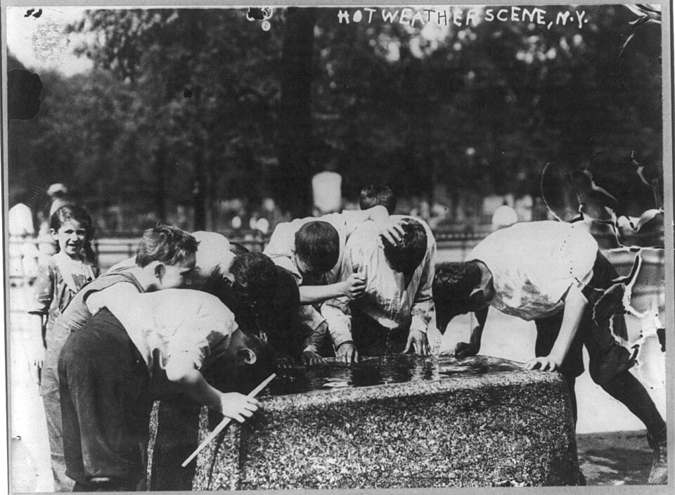 Several children lean over a stone water fountain to drink in a black-and-white photograph captioned “HOT WEATHER SCENE, N.Y.”