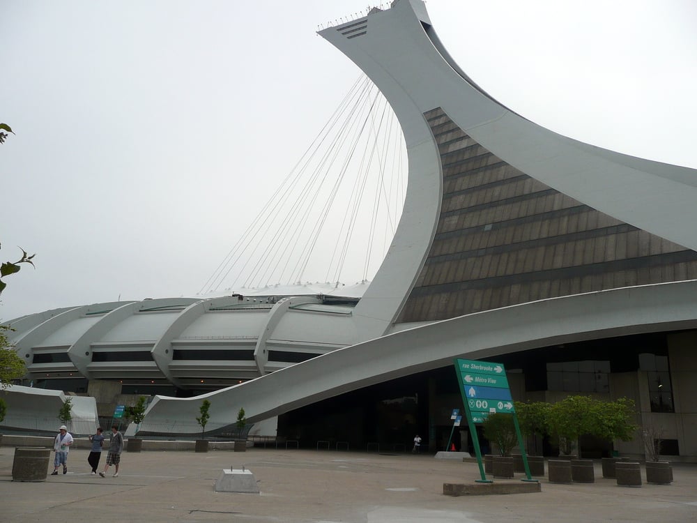 The Montreal Olympic Stadium with its distinctive inclined tower, viewed from a pedestrian plaza with a street sign pointing toward Sherbrooke and Metro Viau.