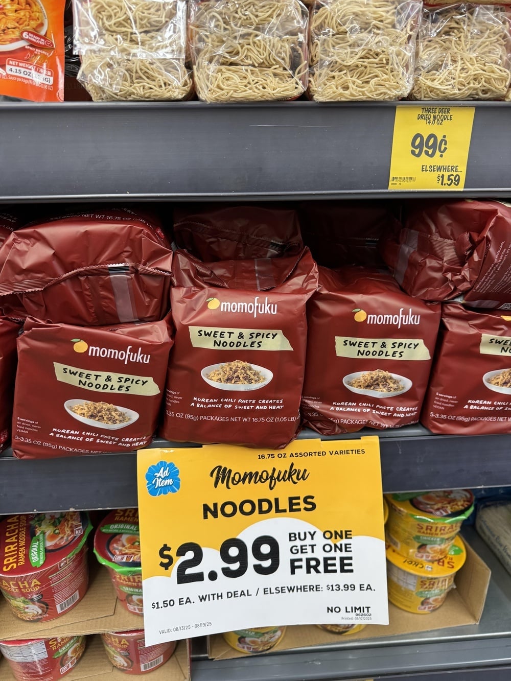 Store shelf displaying Momofuku Sweet & Spicy Noodles in maroon packaging, with a yellow sale sign advertising “Buy One Get One Free” for $2.99.