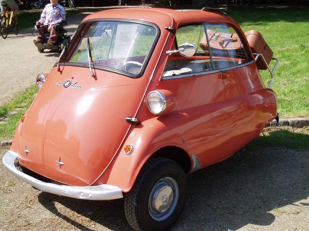 A vintage, coral-colored BMW Isetta bubble car with a luggage case strapped to the rear, parked on a gravel path.