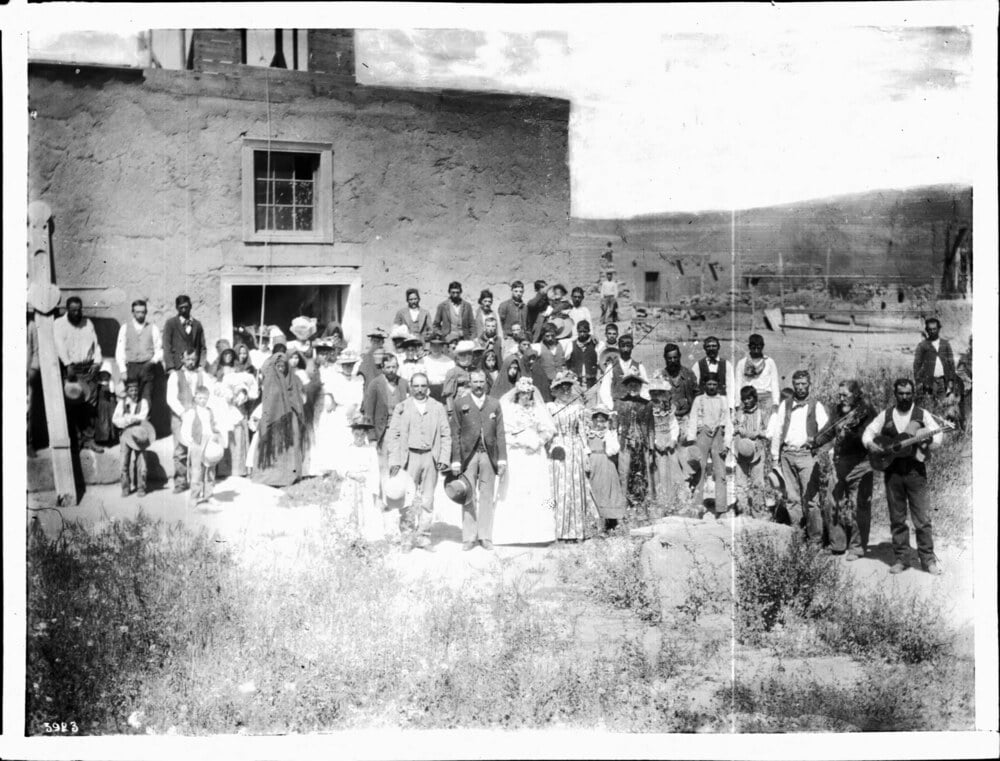 A large group of people in 19th-century attire gathered in front of an adobe building, with a man playing a guitar in the foreground.