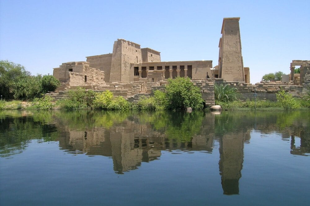 The stone ruins of the Philae Temple complex on an island, reflected in the surrounding river water.