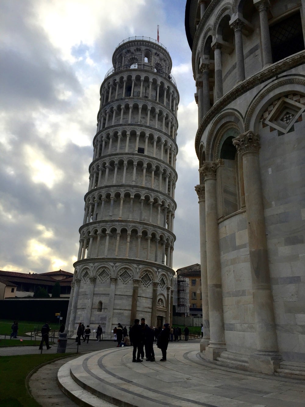 The Leaning Tower of Pisa stands against a cloudy sky with people walking at its base.