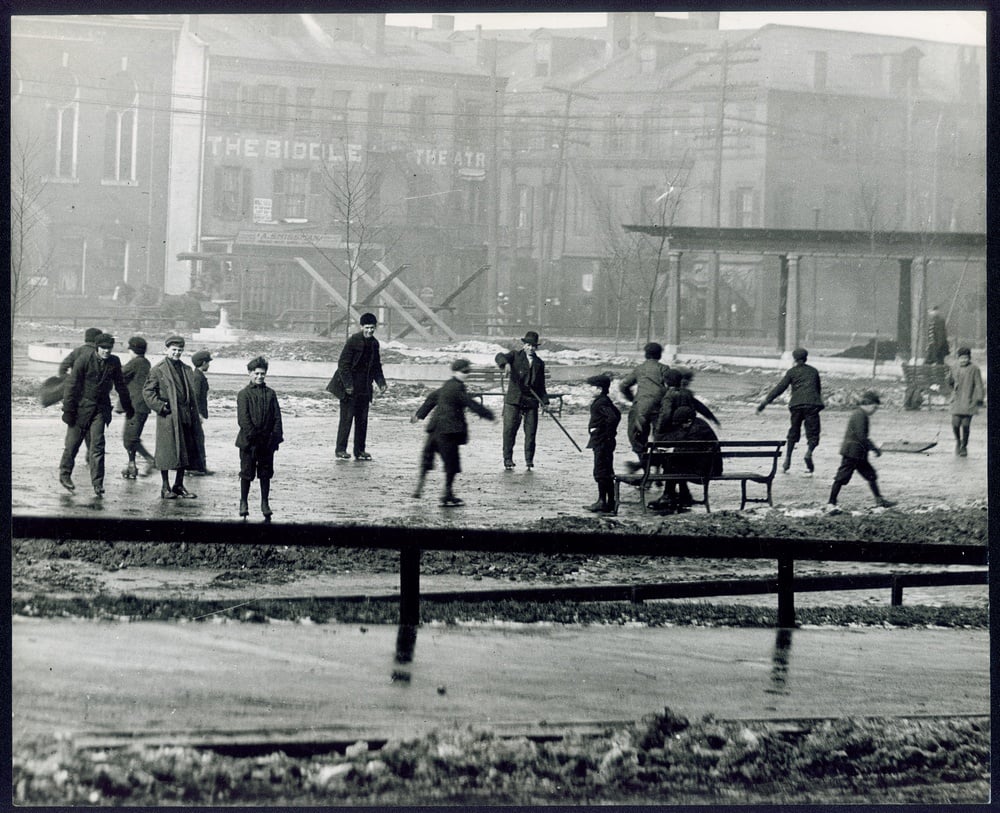 A group of people ice skating on a public pond in front of city buildings.