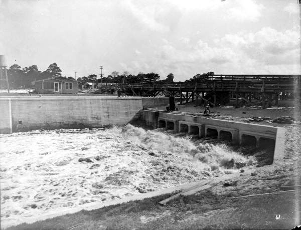 Water rushes through the open floodgates of a concrete dam, with a wooden bridge and buildings visible in the background.