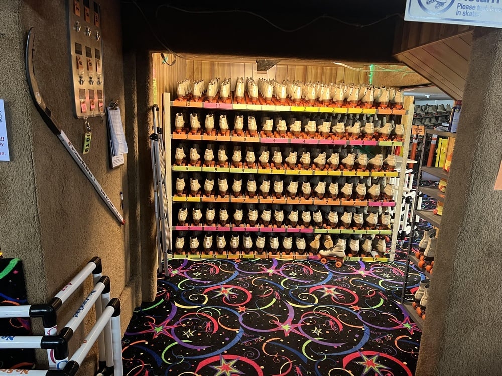 Shelves filled with numerous pairs of roller skates in a rental area with a black patterned carpet.