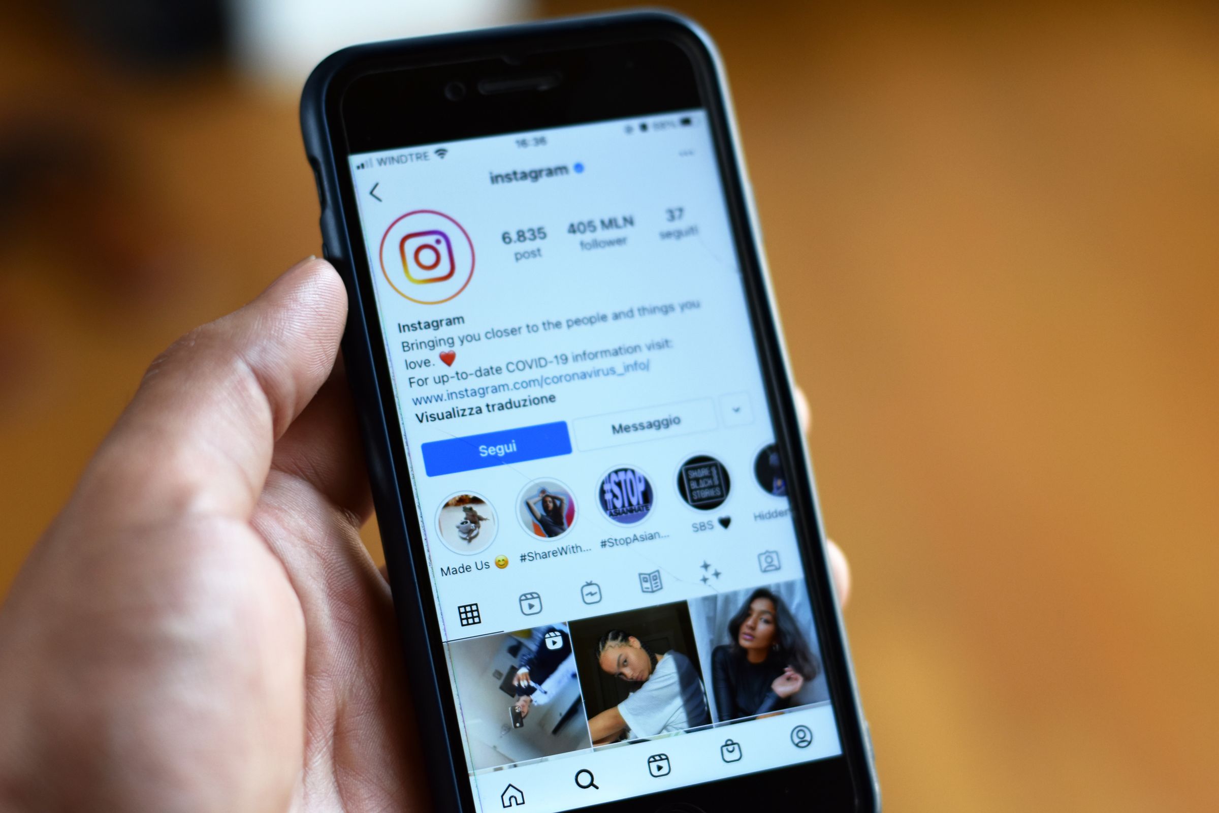 A hand holds a smartphone displaying Instagram’s official profile page, showing its logo, follower count, posts, and profile details. The screen includes posts and highlights at the bottom.